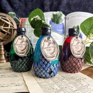 Könnte beinhalten: Drei dekorative Drachenei-Flaschen in Grün, Türkis und Rot. Jede Flasche hat einen strukturierten, schuppenartigen Boden und ein Etikett mit der Aufschrift "Dragon Egg Elixir". Die Flaschen werden auf einem Tisch mit Vintage-Papier und Grünzeug ausgestellt.