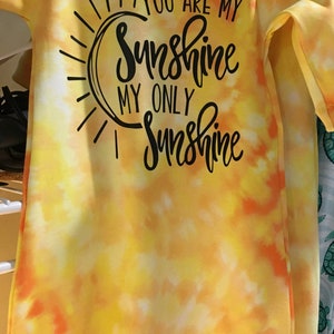 May include: A yellow and orange tie-dye baby onesie with the text "YOU ARE MY Sunshine MY ONLY Sunshine" in black script. The onesie is hanging on a white hanger.