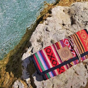 Può includere: Una pochette colorata in tessuto intrecciato con una toppa in pelle che presenta le iniziali "H.C." in un cerchio. La pochette è chiusa e si trova su uno scoglio vicino a un corso d'acqua.