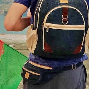 May include: A person wearing a blue shirt and a dark blue backpack with a tan strap and brown leather accents. The person is standing on a rocky surface with a mountain range in the background.