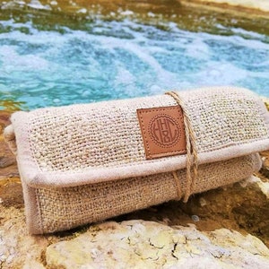 May include: A beige hemp pouch with a brown leather patch featuring the initials "H.H.C." in a circle. The pouch is tied with a brown string and is sitting on a rock near a stream.
