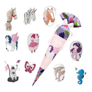 May include: A pink and purple cone-shaped gift bag with the name "Aylin" printed on it, surrounded by embroidered images of a unicorn, ballerina, mermaid, seahorse, butterfly, astronaut, and dolphins. The bag is tied with a white ribbon.