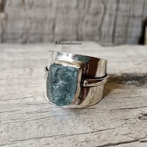 Peut inclure: Une bague en argent avec une grande pierre d'aquamarine bleu clair sertie dans une large bande. La bague est posée sur une surface en bois vieilli.