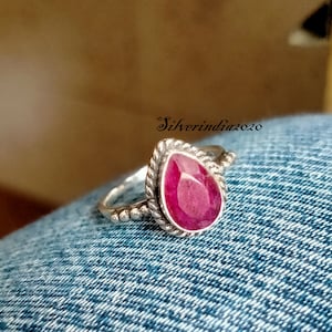 May include: A silver ring with a teardrop-shaped ruby gemstone. The ring has a twisted band with small beads.