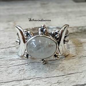 May include: A silver ring with a moonstone and a triple moon design. The ring is set with a large, oval-shaped moonstone in the center, surrounded by three crescent moons.