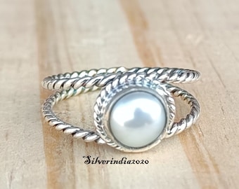 May include: A silver ring with a twisted band and a white pearl set in a silver bezel. The ring is on a wooden surface.