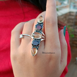 Peut inclure: Une bague en argent avec une rangée verticale de cinq pierres précieuses. Les pierres précieuses sont de couleurs alternées, brun et rouge. La bague est portée sur le doigt d'une personne.