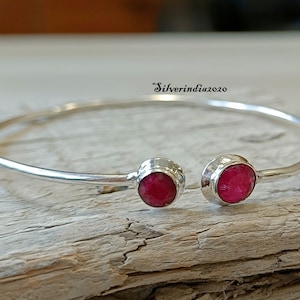May include: A silver bangle bracelet with two red gemstone accents. The bracelet is open and has a simple design.