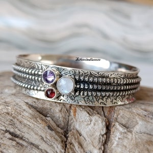 Könnte beinhalten: Silberner Armreif mit drei Edelsteinen: einem violetten Amethyst, einem weißen Mondstein und einem roten Granat. Das Armband hat ein strukturiertes, gemustertes Design.