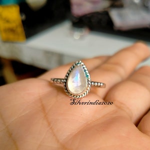 Puede incluir: Un anillo de plata con una piedra de luna en forma de pera engastada en un engaste de cuentas. El anillo está en una mano.