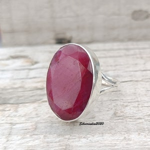 May include: A silver ring featuring a large, oval-cut ruby gemstone. The ruby has a deep red color and is set in a polished silver band. The ring's design is simple, highlighting the beauty of the gemstone.