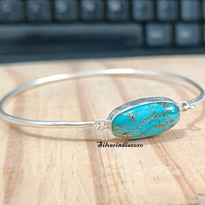 Könnte beinhalten: Ein silbernes Armreif-Armband mit einem türkisfarbenen Stein in der Mitte. Der Stein hat ein marmoriertes Muster aus Gold und Blau.