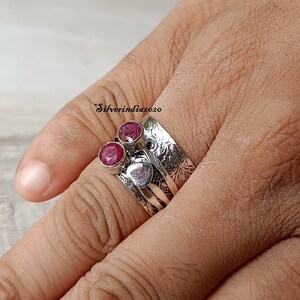 May include: A silver ring with two red gemstones set in the band. The ring has a textured, ornate design.