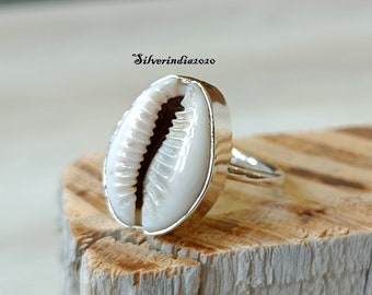 May include: A silver ring with a white seashell set in the center. The ring is on a wooden surface.