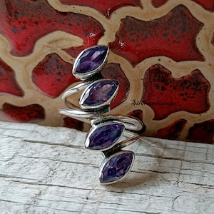 May include: A silver ring with a spiral design and four purple amethyst stones. The ring is on a wooden surface.