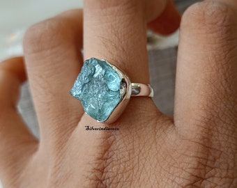 May include: A silver ring with a large, rough-cut aquamarine gemstone. The stone is a light blue color and has a natural, unpolished look. The ring is set in a simple silver band.
