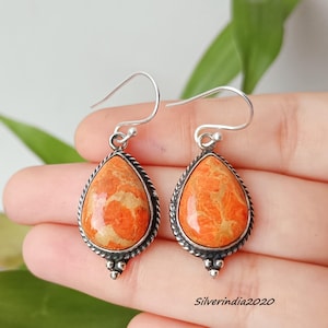 May include: A pair of teardrop-shaped earrings with orange coral-like stones set in a silver-colored metal. The earrings have a textured border and a decorative element at the bottom. The earrings are displayed against a blurred background.