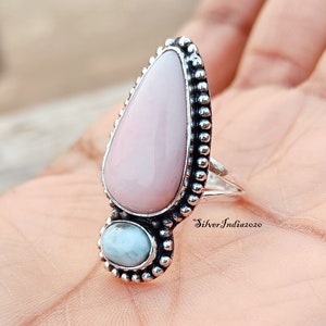 May include: A silver ring with a large pink teardrop-shaped stone and a smaller blue round stone. The ring is set with small silver beads around the stones.