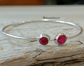 Puede incluir: Un brazalete de plata con dos acentos de piedras preciosas rojas. El brazalete está abierto y tiene un diseño simple.