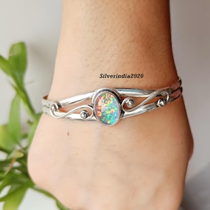Puede incluir: Brazalete de plata con una piedra central ovalada de ópalo iridiscente. La banda de plata presenta un diseño en espiral con pequeños acentos redondos. El ópalo muestra una vibrante gama de colores, incluyendo verde, naranja y azul.