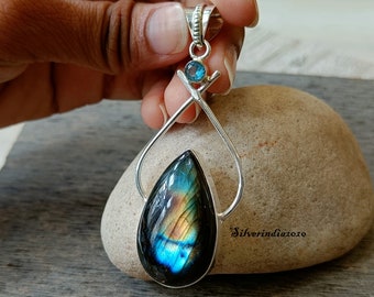 May include: A silver pendant featuring a teardrop-shaped labradorite gemstone with iridescent blue, green, and gold hues. The pendant is accented with a smaller blue gemstone and a silver bail.