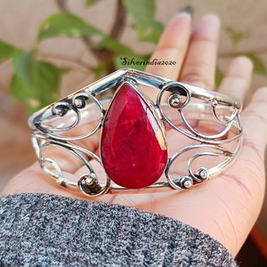 Puede incluir: Un brazalete de plata con una gran piedra preciosa en forma de lágrima de rubí engastada en el centro. El brazalete tiene un diseño delicado y ornamentado con líneas en espiral.
