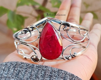 May include: A silver bangle bracelet featuring a large, teardrop-shaped ruby gemstone set in the centre. The bracelet has a delicate, ornate design with swirling lines.