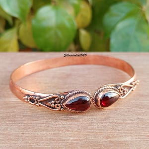 Puede incluir: Un brazalete de cobre con dos acentos de piedras preciosas rojas. El brazalete tiene un diseño adornado con pequeñas cuentas y remolinos.