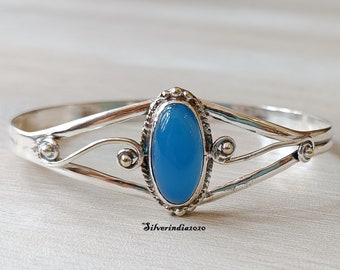 May include: A silver bangle bracelet featuring a light blue oval gemstone set in a silver bezel. The bracelet has a delicate, intricate design with three bands.
