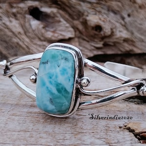 Pode incluir: Uma pulseira de prata com uma gema azul e branca engastada em uma moldura de prata. A pulseira tem um design delicado e torcido.