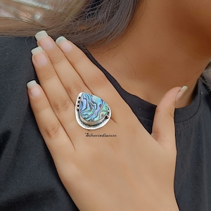 May include: A silver ring with a large iridescent abalone shell set in a teardrop shape. The ring is worn on a person's finger.