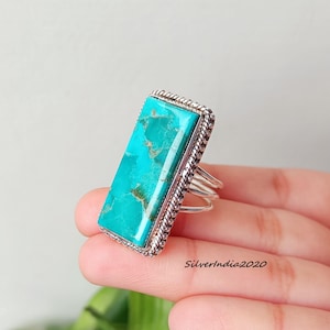 May include: A rectangular turquoise ring with a silver-colored, textured border. The turquoise stone has a mottled pattern of blue and green. The ring has a multi-banded shank. The text "SilverIndia2020" is visible.