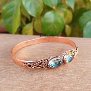 May include: A copper cuff bracelet featuring two teardrop-shaped blue gemstones set at the open ends. The metal band displays intricate twisted wire detailing and small decorative metal beads surrounding the stone settings. Text reads Silverindia2020.