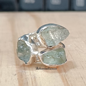 Peut inclure: Une bague en argent ornée de trois pierres précieuses brutes, vert pâle. La bague présente un design unique et organique, avec les pierres serties dans un anneau en argent. Le texte "Silverindia2020" est visible.