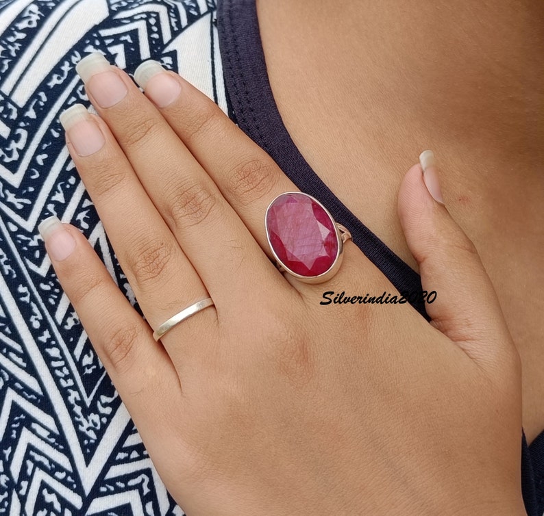 Pode incluir: Um anel de prata com uma grande pedra de rubi oval. O rubi &eacute; de cor vermelha profunda. Um anel de banda de prata simples tamb&eacute;m &eacute; vis&iacute;vel. Os an&eacute;is est&atilde;o em uma m&atilde;o.