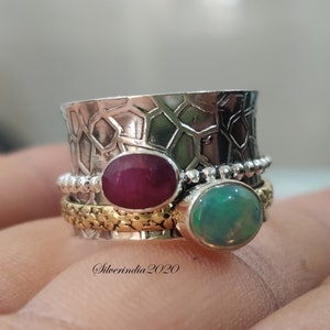 May include: A silver ring with a textured band and two gemstones. The larger gemstone is a green opal, and the smaller gemstone is a red ruby. The ring is on a hand.