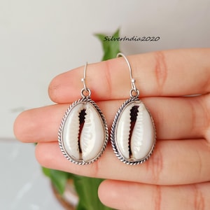 Puede incluir: Pendientes de plata con colgantes en forma de lágrima. Cada colgante presenta una concha blanca con un centro marrón oscuro, encerrada en un marco plateado con un diseño de cuerda trenzada. Los pendientes se sostienen en una mano.