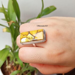 May include: A rectangular ring with a large, yellow and brown stone set in a silver band. The stone has a unique, marbled pattern with shades of yellow, brown, and white. The ring is shown on a hand, with a green plant in the background.