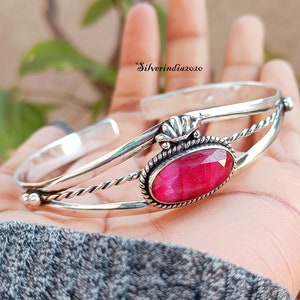 May include: A silver bangle bracelet with a pink gemstone set in a silver oval. The bracelet has a twisted wire design and a decorative silver accent.