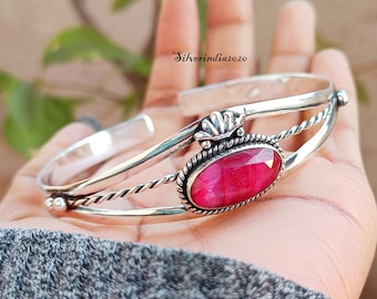 May include: A silver bangle bracelet featuring a pink gemstone set in a silver oval. The bracelet has a twisted wire design and a decorative silver accent.