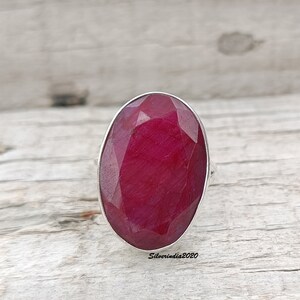 Pode incluir: Um anel com uma grande pedra de rubi oval. O rubi &eacute; de um vermelho profundo e saturado e &eacute; facetado para captar a luz. A crava&ccedil;&atilde;o do anel &eacute; prateada e a pedra preciosa &eacute; o ponto focal. O anel est&aacute; sobre uma superf&iacute;cie de madeira clara.