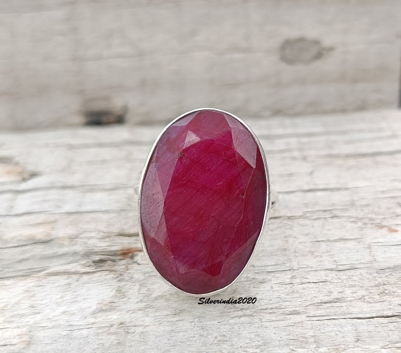 Pode incluir: Um anel com uma grande pedra de rubi oval. O rubi &eacute; de um vermelho profundo e saturado e &eacute; facetado para captar a luz. A crava&ccedil;&atilde;o do anel &eacute; prateada e a pedra preciosa &eacute; o ponto focal. O anel est&aacute; sobre uma superf&iacute;cie de madeira clara.