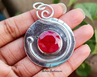 May include: A silver pendant with a large, faceted ruby gemstone at its center. The pendant has a circular design with a spiral at the top and a curved line detail. The text "SilverIndia2020" is visible on the bottom right.