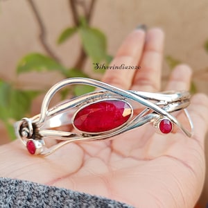 May include: A silver bangle bracelet with a large oval ruby gemstone and two smaller ruby gemstones. The bracelet has a delicate, intricate design.
