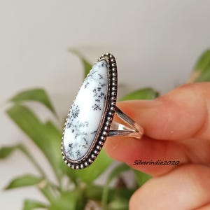 May include: A silver ring featuring a large, oval-shaped white stone with black dendritic inclusions. The stone is framed by a beaded silver border. The ring's band is split. The ring is held up by a hand.