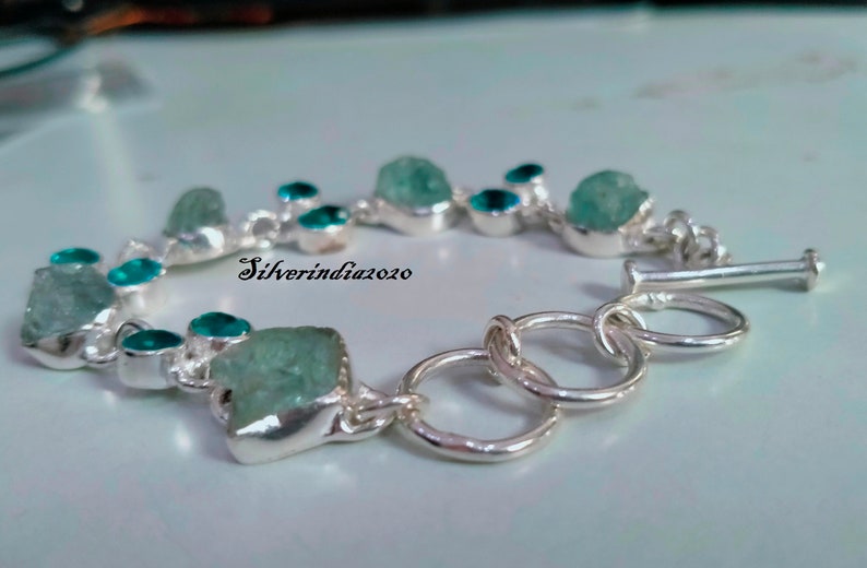 Puede incluir: Un brazalete de plata con cierre de broche presenta una fila de piedras preciosas azul claro y turquesa. El brazalete est&aacute; hecho con una cadena de plata y tiene un broche de plata.