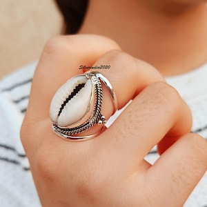 Op de afbeelding: Een zilveren ring met een wit zeeschelp-middelpunt. De ring heeft een gedetailleerd ontwerp met gevlochten en kralenaccenten. De zeeschelp is ovaal van vorm met een donker, getextureerd interieur. De ring wordt aan een vinger gedragen.