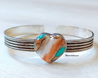 May include: A silver cuff bracelet with a heart-shaped turquoise stone. The stone has a white, orange, and turquoise pattern. The bracelet has three silver bands with a rope design.