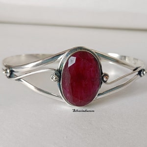 Puede incluir: Un brazalete de plata con una gran piedra preciosa roja de forma ovalada en el centro. El brazalete tiene un diseño delicado y retorcido.