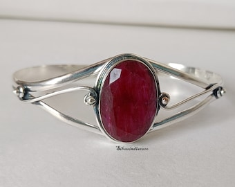 Puede incluir: Un brazalete de plata con una gran piedra preciosa roja de forma ovalada en el centro. El brazalete tiene un diseño delicado y retorcido.